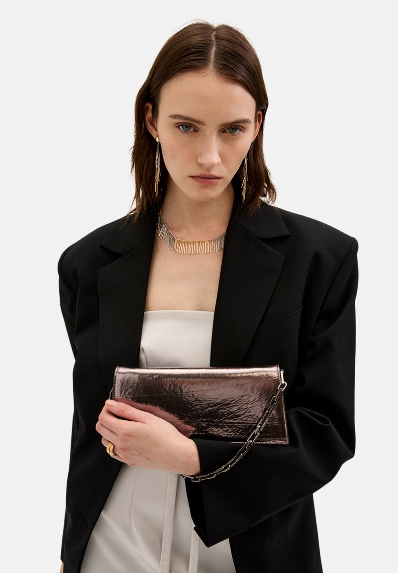 Pochette in metallo con finitura lucida, forma rettangolare, tracolla staccabile, tenuta in mano su uno sfondo di blazer nero.
