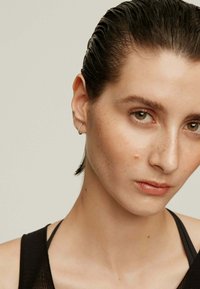 Pendiente de aro plateado, modelo con cabello slicked-back, tono de piel neutro, maquillaje minimalista, vistiendo una blusa negra con tirantes visibles.