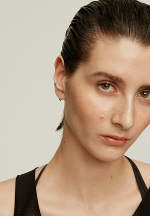 Pendiente de aro plateado, modelo con cabello slicked-back, tono de piel neutro, maquillaje minimalista, vistiendo una blusa negra con tirantes visibles.