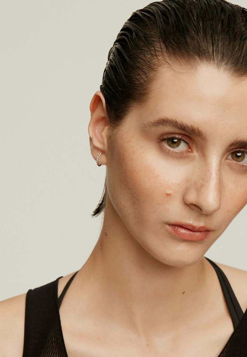 Pendiente de aro plateado, modelo con cabello slicked-back, tono de piel neutro, maquillaje minimalista, vistiendo una blusa negra con tirantes visibles.