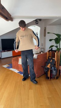Un homme portant un pull beige avec des rayures noires sur les manches se tient dans un salon en tirant le pull vers l'extérieur.
