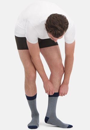 Mann, der sich vorbeugt, schwarze Shorts und weißes T-Shirt trägt, blaue und weiße gestreifte Socken vor einem weißen Hintergrund hochzieht.