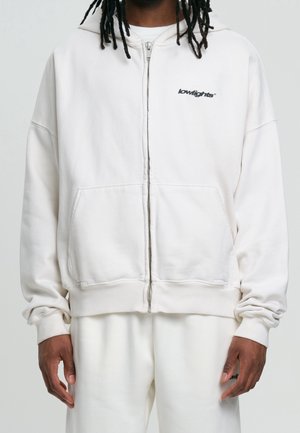 Man draagt een witte hoodie met ritssluiting met het logo "lowlights" op de borst en bijpassende witte broek tegen een neutrale achtergrond.
