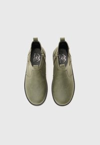Zapatos slip-on verde oliva hechos de un material suave, con paneles laterales elásticos, punta redondeada y suela negra en contraste.
