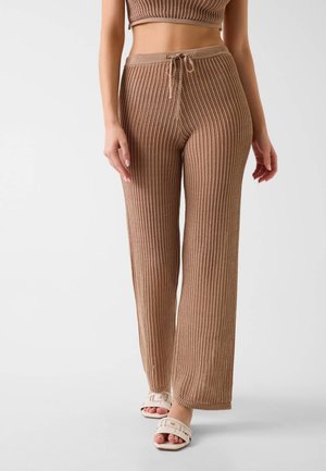 Femme portant un pantalon en maille marron avec une taille à cordon et des sandales blanches ouvertes aux orteils, debout devant un fond clair uni.