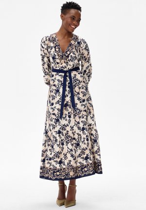 Uśmiechnięta osoba w długiej kremowej sukience z ciemnoniebieskimi wzorami kwiatowymi i pasującym paskiem, stojąca z rękami za plecami.