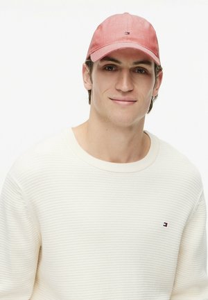 Joven con un jersey de punto blanco y una gorra de béisbol roja desgastada con un pequeño logo, sonriendo contra un fondo blanco liso.