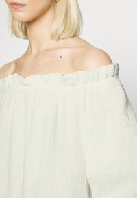 Top cinza claro, com decote off-the-shoulder, feito de um tecido texturizado e amarrotado, com um acabamento de babados na gola. O material parece suave e leve.