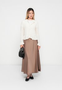 Frau in einem weißen strukturierten Cardigan und einem langen braunen Rock, die eine schwarze Handtasche hält und vor einem schlichten weißen Hintergrund steht.