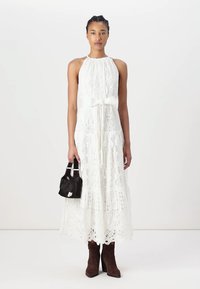 Robe en dentelle blanche avec un col halter, ornée de motifs complexes et d'un nœud à la taille. Associée à des bottes en suède marron et un sac à main noir.