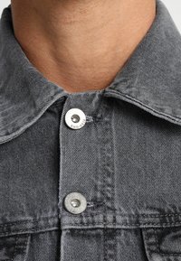 Harmaa denim-takki, jossa on lähellä oleva näkymä kauluksesta, varustettuna metallisilla painonapeilla ja teksturoiduilla kangasyleillä.