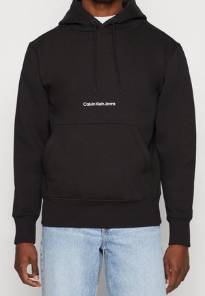 Muž v černé mikině Calvin Klein Jeans s přední kapsou a světle modrých džínách, stojící před jednobarevným pozadím.