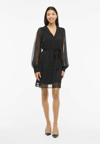 Czarna sukienka w białe groszki z przezroczystymi długimi rękawami, dekoltem w serek i paskiem w talii; posiada długość do kolan i zwiewną fakturę.