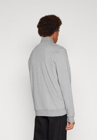 Felpa grigia con collo alto, realizzata in tessuto morbido, con maniche lunghe e orlo a coste, indossata con pantaloni neri.