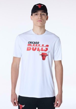 Young man wearing a white Chicago Bulls t-shirt and black cap, both featuring the team's red bull logo. Black shorts with small red bull logo.