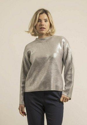 Pull en tricot argenté avec une texture côtelée, des manches longues et un col montant. Porté avec un pantalon bleu marine, il présente une finition brillante.