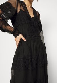 Femme portant une robe en dentelle noire transparente avec motif à pois, manches longues et bordure festonnée, main posée sur la hanche sur fond blanc.