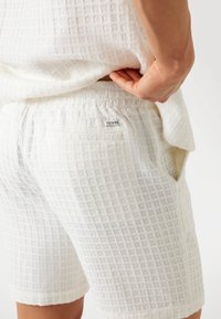 White shorts in a textured, grid-like material with elastic waistband and side pockets. Features a small "GUESS" label on the back.