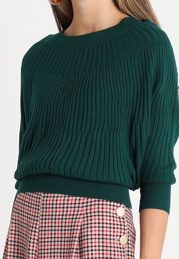 Femme portant un pull côtelé vert foncé à manches trois-quarts et une jupe à carreaux roses avec deux boutons métalliques sur le côté.