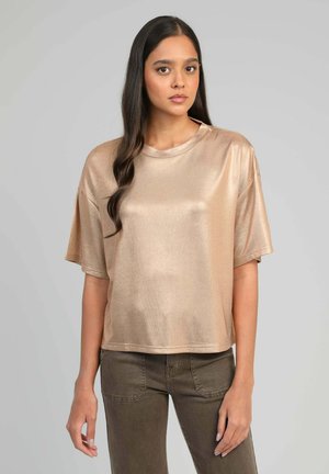 Femme aux longs cheveux foncés portant un haut beige brillant à manches courtes et un pantalon vert olive, debout devant un fond gris uni.