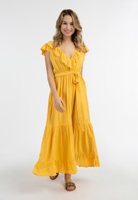 Rochie maxi galbenă cu mâneci volan, decolteu în V și talie legată. Prezintă fustă în straturi și țesătură ușoară, texturată. Purtată cu sandale decorative.