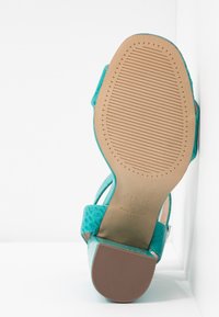 Sandale à talon haut turquoise montrant une semelle en caoutchouc texturée avec motif antidérapant et talon bloc sur fond blanc.