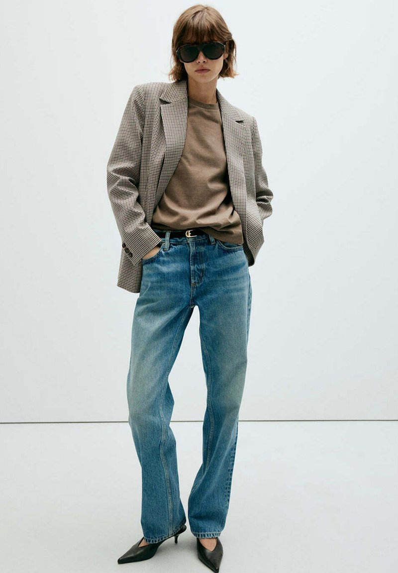 Femme portant de grandes lunettes de soleil foncées, un t-shirt beige, un blazer à carreaux, un jean bleu ample et des talons noirs pointus, debout les mains dans les poches.