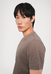 Brown short-sleeve t-shirt featuring a repeating "MK" pattern, round neckline, and a smooth texture. The design is fitted and modern.