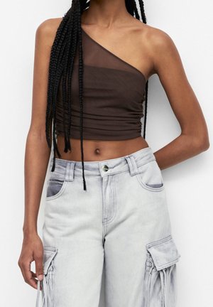 Mujer con largas trenzas que lleva un top corto de un solo hombro, transparente, de color marrón oscuro, y unos vaqueros cargo de talle alto en gris claro con bolsillos.