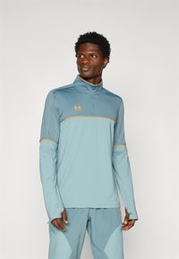 Camiseta deportiva de manga larga azul claro con media cremallera, detalles en naranja y un cuello alto. Presenta mangas texturizadas y un diseño ajustado.