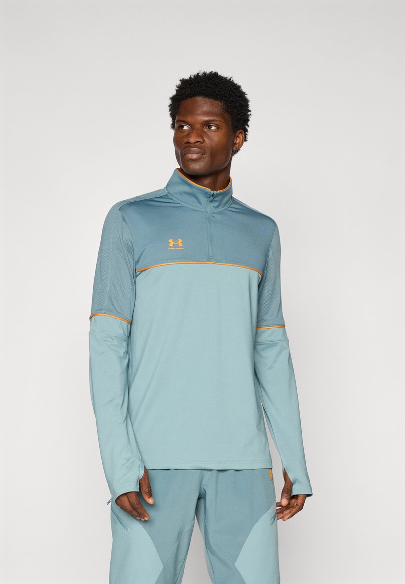 Camiseta deportiva de manga larga azul claro con media cremallera, detalles en naranja y un cuello alto. Presenta mangas texturizadas y un diseño ajustado.