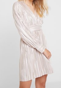 Robe plissée en rose doux avec des rayures métalliques scintillantes, décolleté en V profond et manches longues, dotée d'une taille cintrée pour une silhouette structurée.