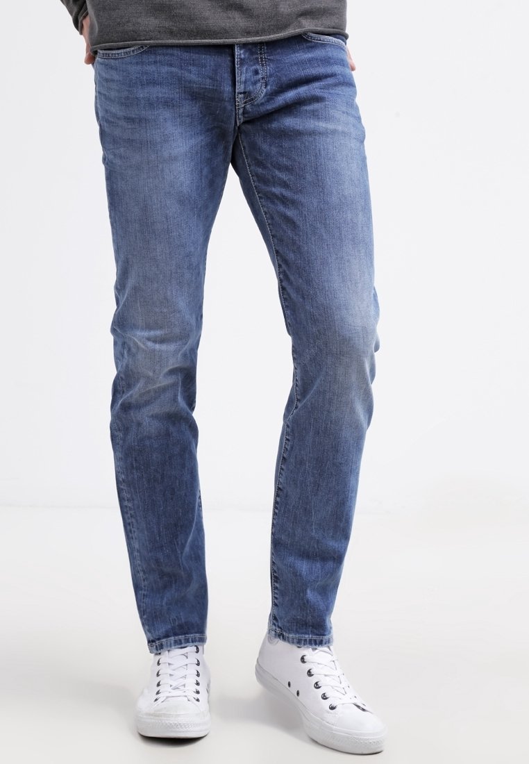 Světlé modré džínsy s úzkým střihem, které mají klasický pět-pocket design a jemné vrásečky na stehnech, doplněné bílými teniskami.