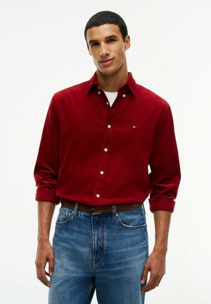 Mann trägt ein rotes Hemd mit Knöpfen über einem weißen Unterhemd, blaue Jeans und einen braunen Gürtel vor einem einfachen weißen Hintergrund.