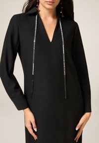 Zwarte jurk met lange mouwen, een kraag en een V-hals, voorzien van sierstenen en satijnen lintaccenten langs de voorkant.