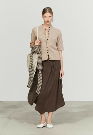 Donna che indossa un cardigan beige abbottonato, gonna lunga marrone scuro, scarpe bianche, che tiene una borsa beige con una sciarpa a righe sulla spalla.