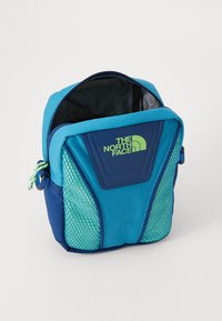 Saco de tecido azul com detalhes em malha e um logotipo frontal. Possui uma abertura com zipper. O design inclui uma área texturizada e cores contrastantes.