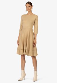 Abito beige alla lunghezza del ginocchio con corsetto aderente, maniche a tre quarti, gonna plissettata e dettagli a taglio orizzontale vicino all'orlo.