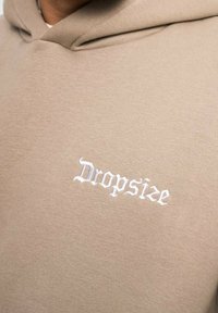 Sudadera beige con una textura suave que presenta bordado blanco con el texto "Dropsize" en estilo gótico. El diseño incluye una capucha con cordones.