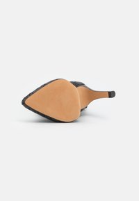 Zapato de tacón alto con punta afilada, parte superior negra texturizada y suela beige lisa. Presenta intrincados patrones de corte en los laterales.