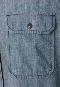 Modrá džínsová košile s kapsou na klopu, která je zajištěna černým knoflíkem s vyrytým nápisem "Replay 1981" uprostřed.