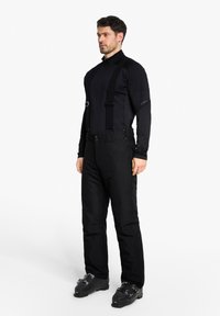 Mann in schwarzen Skihosen mit Hosenträgern, schwarzem Langarmshirt und schwarzen Skischuhen, der vor einem schlichten weißen Hintergrund steht.