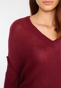 Maglione in tricot bordeaux con scollo a V, vestibilità ampia e tasca laterale. La texture è leggera e leggermente trasparente.