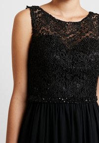 Ärmlös svart klänning med transparent spets på överdelen, blommönsterbroderier, små pärldekorationer och detaljerade bälten med pärlor.