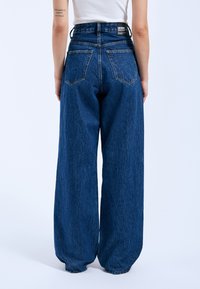 Hoch taillierte, weite Jeans in dunkelblauem Denim. Mit zwei Gesäßtaschen, sichtbaren Nähten und einem Etikett am Bund.