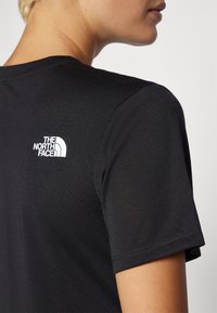 Črna bombažna majica s kratkimi rokavi, ki ima belo logo "The North Face" na zgornjem hrbtu blizu izreza.
