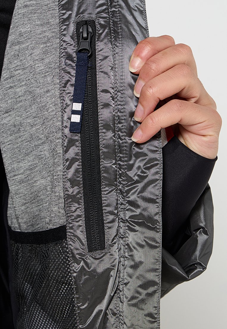 Chaqueta deportiva gris con un bolsillo con cremallera, acabada en textura brillante y con tejido de malla contrastante a los lados. Acentos en negro y azul marino.