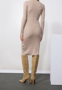 Femme portant une robe en maille côtelée beige avec des poignets boutonnés, associée à des bottes à talons hauteur genou en daim marron clair, debout devant un fond uni.