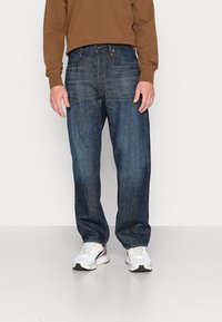 Mörkblå jeans i rak modell med subtila slitningseffekter. Kombinerade med en brun långärmad tröja och vita sportskor.