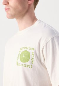 White cotton t-shirt featuring green circular graphic with smiley face and text: "Scotch & Soda Amsterdam Art Dept Class of '23." Short-sleeved.